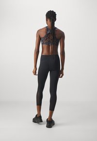 Kvinna med långa flätor som bär en svart mönstrad sport-bh, svarta korta leggings och svarta sneakers, stående vänd bortåt på en grå bakgrund.