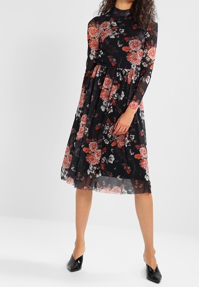 Femme portant une robe midi noire transparente à motifs floraux, manches longues et taille cintrée, assortie à des escarpins noirs à bouts pointus.