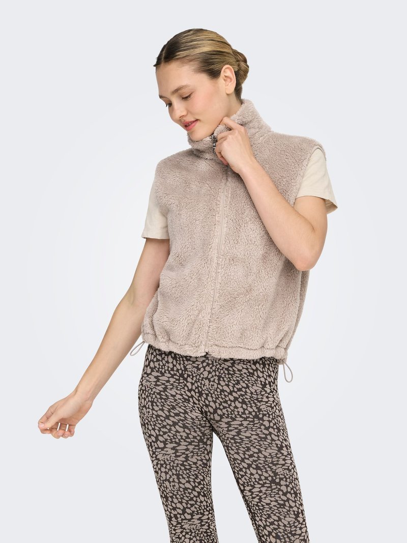 Gilet en fausse fourrure beige clair avec un col montant et une fermeture éclair. Associé à des leggings foncés à motifs tachetés.