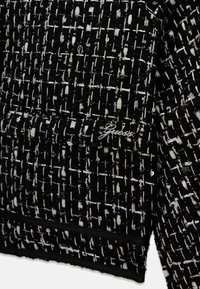 Tkanina tweedowa w czerni i bieli z teksturowanym wzorem, z subtelnym detalem logo. Strukturalny kształt i kontrastowe akcenty wykończenia.
