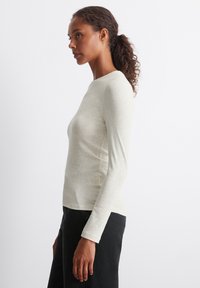 Lys beige langermet topp med rund hals og kroppsnær design. Stoffet ser mykt og litt teksturert ut, kombinert med mørke bukser.