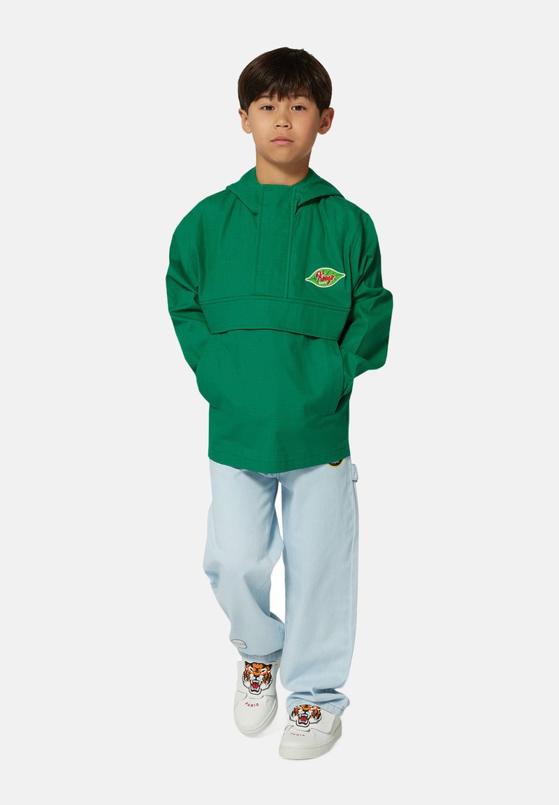 Pullover verde leggero con cappuccio, dotato di tasca frontale e dettagli con logo; abbinato a jeans baggy blu chiaro e scarpe bianche con un design di tigre.