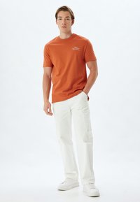 Orange kortærmet t-shirt med rund hals, prydet med hvid tekst. Sammen med hvide cargo bukser og hvide sneakers. Enkel baggrund.