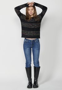 Fekete rövidített pulóver színes vízszintes csíkokkal, kék, skinny farmerral és térd fölé érő fekete csizmával párosítva. Az anyagok sima textúrájúak.