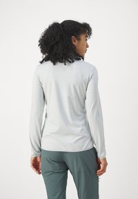 Lichtgrijs langemouwen shirt met een gladde textuur, aangesloten silhouet en ronde onderrand. Graagd met donkergroene broek, zichtbaar vanuit een zijaanzicht.