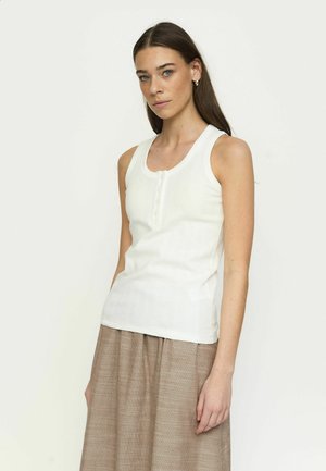 Jonge vrouw met lang bruin haar, gekleed in een wit mouwloos topje met knopen en een beige geruite rok, staand tegen een effen lichte achtergrond.