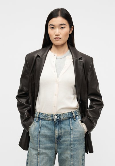 Νέα γυναίκα με μακριά σκούρα μαλλιά φορά ελαφριά πουκάμισα σε στρώσεις με κουμπιά, υπερμεγέθη σκούρο δερμάτινο σακάκι και τζιν ψηλόμεσα μπλε, με τα χέρια στις τσέπες.