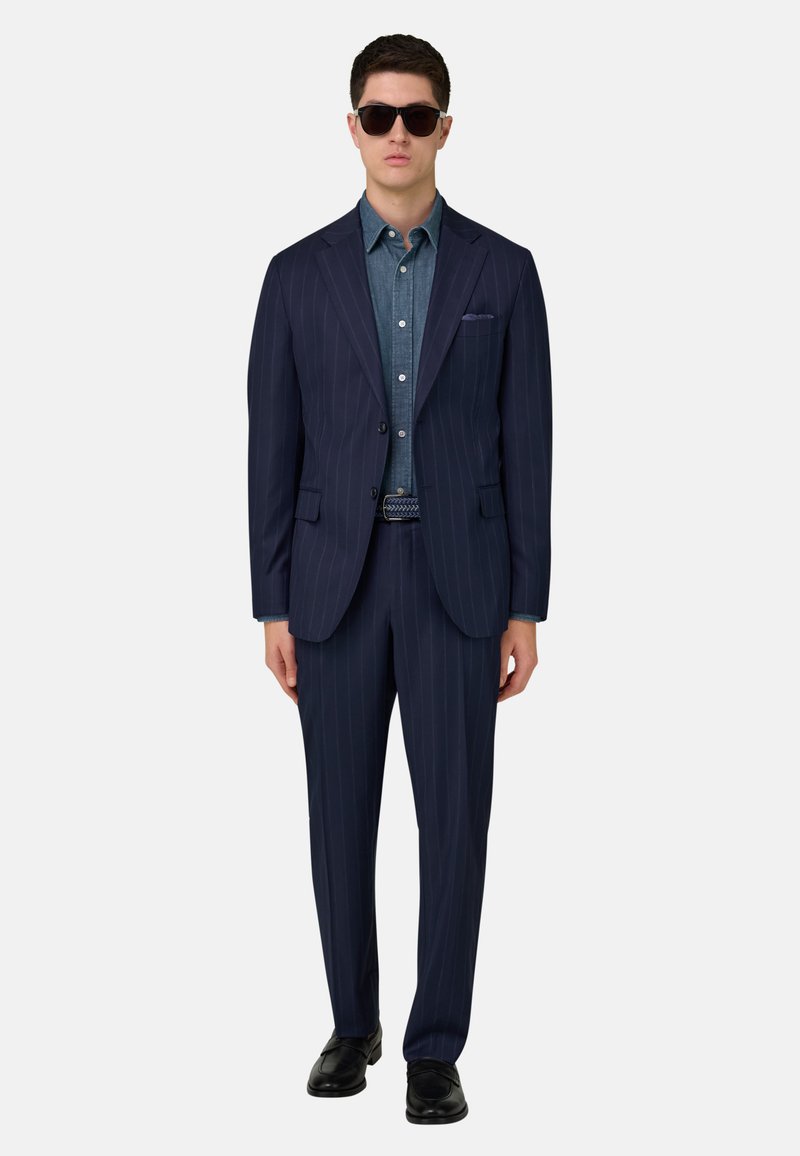 Homme portant un costume bleu marine à fines rayures, une chemise bleu clair à boutons, une ceinture noire, des mocassins noirs et des lunettes de soleil noires, debout devant un fond blanc.