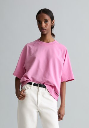 Tânăr cu păr închis la culoare prins într-o coadă joasă, purtând o cămașă roz supradimensionată cu mâneci scurte, băgată într-o pereche de pantaloni albi, cu o curea neagră.