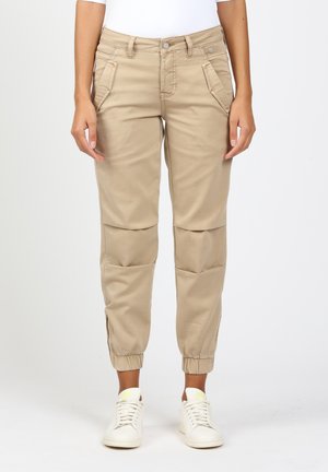 Hiekanväriset jogger-housut, joissa on joustavat lahkeet, seisovan henkilön yllä, yhdistettynä valkoisiin lenkkareihin ja valkoiseen yläosaan.