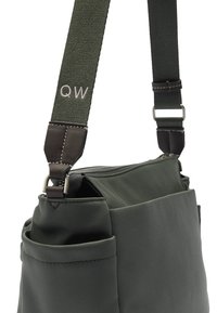 Zielona torba materiałowa z zamkiem błyskawicznym, skórzanymi akcentami, regulowanym paskiem i zewnętrzną kieszenią. Wzór haftowany "QW" na pasku.