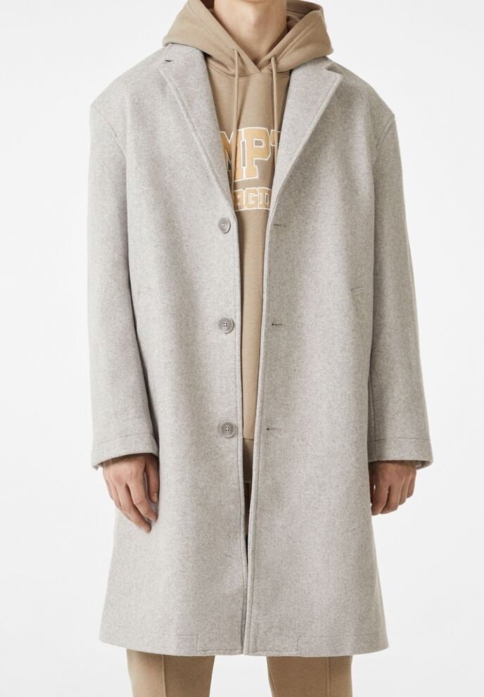 Hellgrauer Wollmantel mit Reverskragen, einzelner Knopfschluss und Seitentaschen, übereinander getragen mit einem Beigen Hoodie.