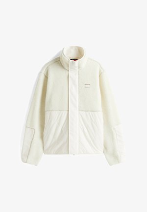 Chaqueta de forro polar blanca con panel inferior acolchado, cuello alto y un pequeño parche con el logo en el pecho. Textura suave, corte relajado y mangas largas.