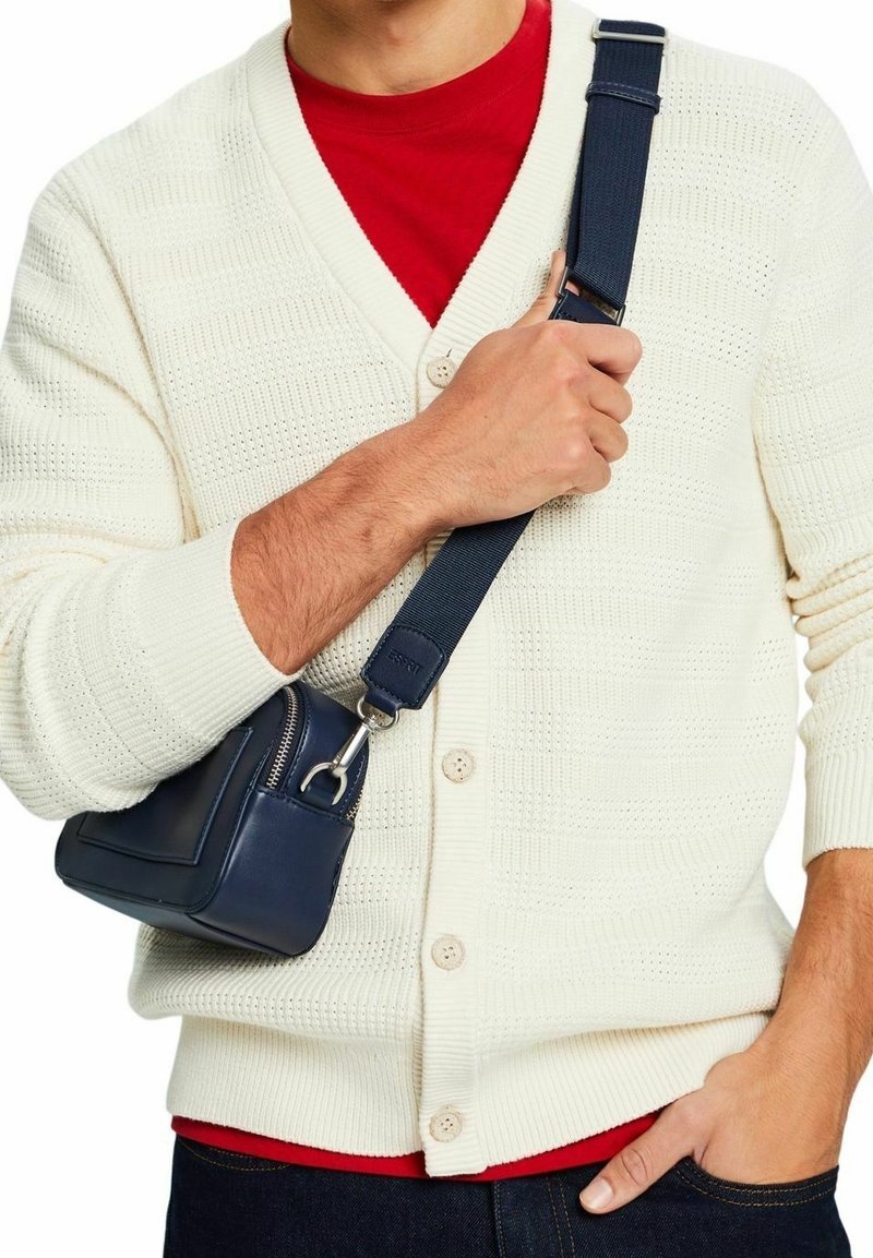 Homme portant un cardigan en maille blanc et une chemise rouge tient un sac bandoulière bleu marine par sa sangle devant la poitrine.