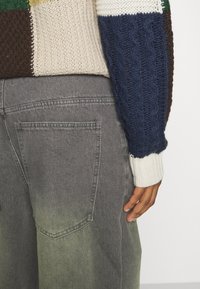 Suéter de punto con secciones en verde, amarillo, marrón, crema y azul; presenta una textura de punto de cable. Llevado con pantalones de mezclilla gris oscuro, ligeramente desgastados.