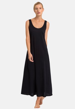 Kobieta stoi boso, ubrana w długą, czarną sukienkę bez rękawów z okrągłym dekoltem i małą kieszonką na piersi.