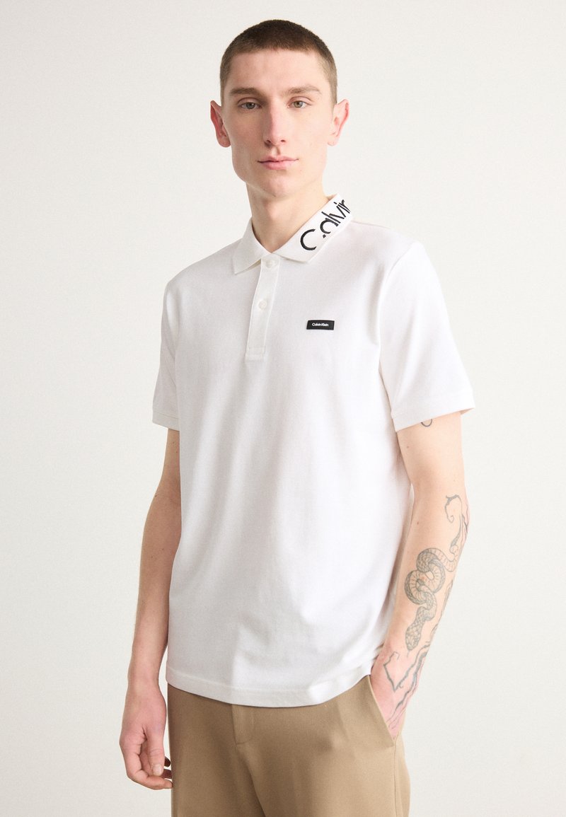 Witte poloshirt met korte mouwen, voorzien van een zwart geborduurd logo op de kraag en een klein zwart label op de borst. Zacht stof.