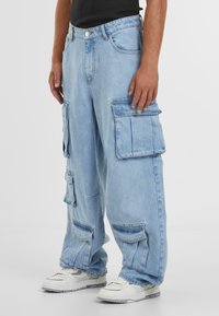 Lyseblå cargo-jeans laget av denim, med flere lommer og løs passform. Brukt med hvite joggesko.