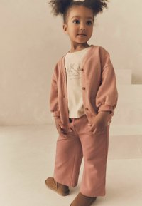 Barn i en pink cardigan og matchende bukser, lagdelt over en lys skjorte med et kaninprint. Med bløde teksturer og brune støvler.