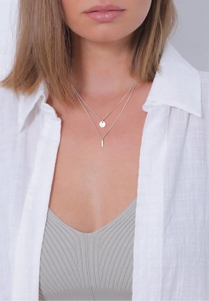 Femme aux cheveux châtain clair portant une chemise blanche ouverte, un haut gris à côtes et deux colliers argentés superposés avec des pendentifs ronds et en barre.
