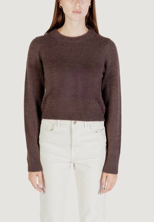 Donna che indossa un maglione lungo a maniche lunghe, morbido di colore marrone scuro e pantaloni a vita alta color crema, in piedi con le braccia rilassate lungo i fianchi.