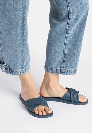 Blue slide sandals with woven detailing and a flat sole, paired with light blue cuffed denim jeans. Smooth rubber material and casual design.