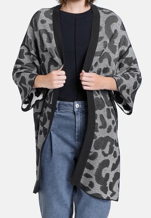 Frau trägt einen schwarz-grauen Cardigan mit Tiermuster und offenem Vorderteil über einem schwarzen Top und blauen hoch taillierten Jeans, hält die Ränder des Cardigans.