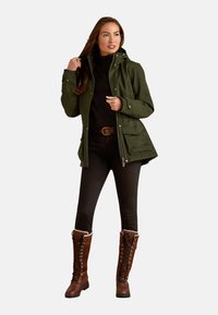 Groene, gewatteerde jas met een gestructureerde afwerking en voorzakken, gecombineerd met zwarte leggings en bruine kniehoge veterschoenen.
