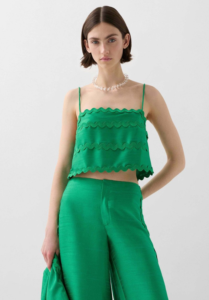 Grøn cropped top med bølgede kanter, parret med matchende grønne bukser med bred ben. Glat tekstur, minimalistisk design og justerbare stropper.