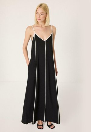 Vrouw draagt een zwarte maxi-jurk met dunne bandjes en witte verticale biezen, gecombineerd met zwarte open hakken, staand tegen een effen achtergrond.