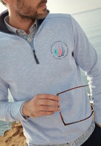 Lichtblauwe kwart-rits pullover met textuur en subtiel patroon, voorzien van een logo met kleurrijke zeilboot graphics; gelaagd met een gestreept shirt.
