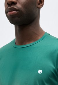 Κοντινό πλάνο ενός άνδρα που φοράει πράσινο μπλουζάκι με στρογγυλή λαιμόκοψη και μικρό λευκό κυκλικό λογότυπο στο στήθος, με απλό φόντο.