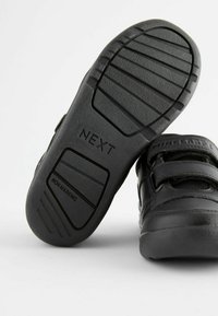 Chaussures noires pour enfants avec une semelle en caoutchouc texturé, des bandes Velcro et une surface supérieure lisse. Présente les inscriptions "NON MARQUANT" et "NEXT".