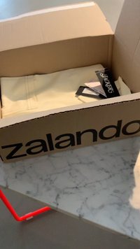 Cardboard box labeled "zalando" contains folded cream-colored pants with visible stitching and tags. Set on a marble surface with red accents.
