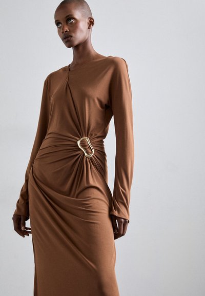 Vestido largo de jersey marrón con un detalle retorcido en la cintura, que presenta un acento dorado. Las mangas son ajustadas y la tela parece suave.