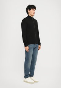 Mann steht, trägt einen schwarzen Reißverschlusspullover, blaue Jeans und weiße Sneakers vor einem einfarbigen hellen Hintergrund.