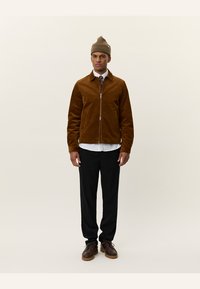 Brun kordjangjacka med dragkedja, bärs över en vit skjorta och ett mönstrat slips. Svarta byxor och bruna skor kompletterar outfiten.