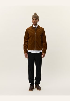 Chaqueta de pana marrón con cremallera frontal, usada sobre una camisa blanca y una corbata con estampado. Pantalones negros y zapatos marrones completan el atuendo.