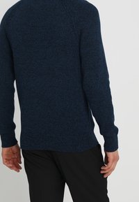 Man med en mörkblå strukturerad långärmad tröja och svarta byxor, sedd bakifrån mot en enfärgad bakgrund.