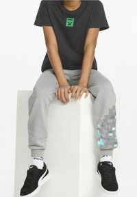 Persona con una maglietta nera Puma e pantaloni grigi con un design pixelato di Minecraft, seduta su un blocco bianco, con le mani appoggiate davanti.