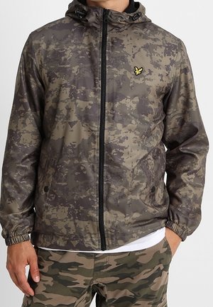 Mand iført en brun og grøn digital camo hættejakke med sort lynlås og matchende camo bukser, stående med den ene hånd i lommen.