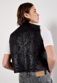 Donna con i capelli corti castani indossa un gilet nero a motivi sopra una maglietta bianca e jeans grigi, rivolta di spalle e mentre guarda di lato.