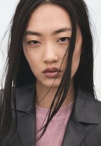 Portrait en gros plan avec de longs cheveux noirs, portant une veste en cuir noire sur un pull rose clair. Éclairage doux avec un fond neutre.