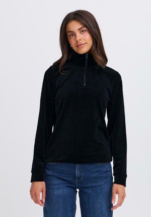 Pull en velours côtelé noir avec col montant, fermeture éclair frontale à mi-hauteur, manches longues et un motif côtelé texturé. Assorti à un jean bleu.