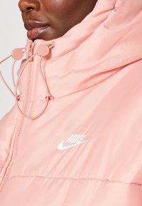 Rosa puffjacka med hög krage, utrustad med dragsko och en vit Nike-logotyp. Materialet verkar slätt och blanka.