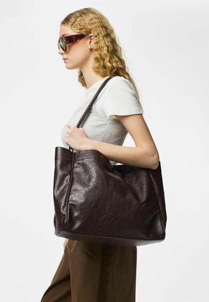 Kvinne med bølgete blondt hår bærer en stor mørkebrun skinnveske over skulderen, iført hvit kortermet topp og solbriller.