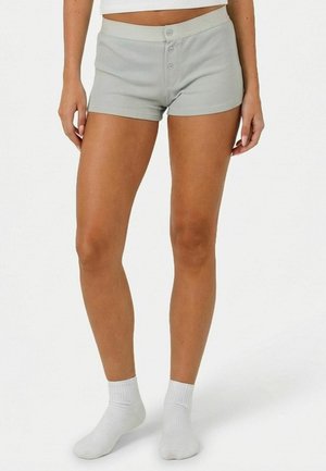 Ženské nohy v svetlosivých šortkách s gombíkmi vpredu a bielych členkových ponožkách, stojace na bielom pozadí.