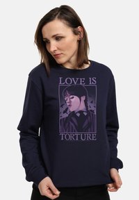Donna che indossa una felpa blu navy con una grafica viola di una ragazza con trecce e pipistrelli, accompagnata dal testo "L'AMORE È TORTURA."