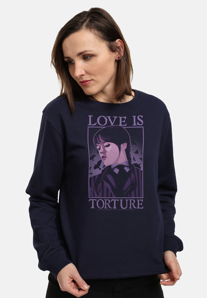 Donna che indossa una felpa blu navy con una grafica viola di una ragazza con trecce e pipistrelli, accompagnata dal testo "L'AMORE È TORTURA."