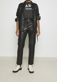 Chaqueta de camisa de cuero negra sobre una camiseta gráfica negra, combinada con pantalones de cuero negros y botas de tobillo blancas con suelas negras.